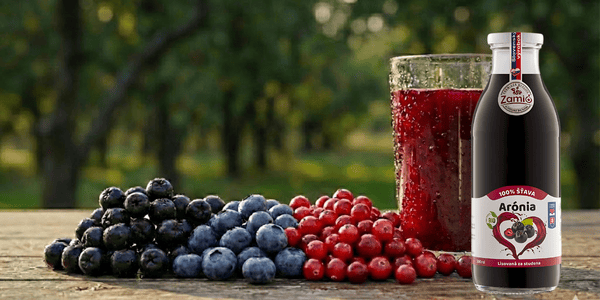 na obrazku je drevený stol a pozadie v zahrade , na stole je oocie bobule a flasa aronie od zamia a pohar zo šťavov